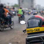 Um capacete de moto‑táxi pousado no assento de uma motorizada, em primeiro plano, com kupapatas a circular ao fundo numa rua movimentada, junto a um mercado urbano. A cena retrata a dinâmica da mobilidade informal em Luanda, com motociclistas e vendedores ao longo da via.